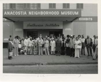 A group of people stand outside of a theater named the Anacostia Neighborhood Museum. They are standing on a sidewalk. 