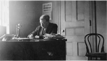 A man sits at his desk, writing. An empty chair is also in the frame.