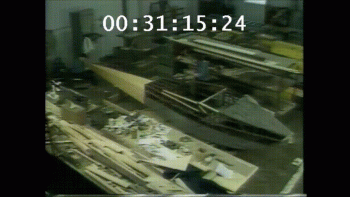 Two men in blue coveralls working on a wooden plane in a warehouse.