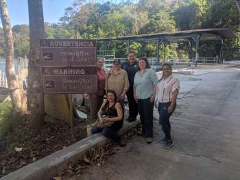 Six people smiles for a photograph near a sign that reads, "Warning Crocodiles in the Area" in English and Spanish. Behind them is a forest of trees and a boat dock.