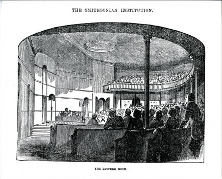 Lecture Hall looking toward the stage. A man is demonstrating an experiment to an audience seated in