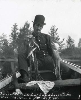 A man in a suit and hat stares directly at the camera, holding a fish and a net. Trees and a body of