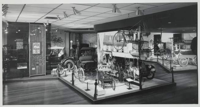 Carriages, automobiles, and cycles on display in an exhibit hall. 