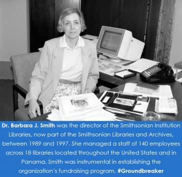 Smith sits at a desk in front of a pile of papers and an older-looking computer. 