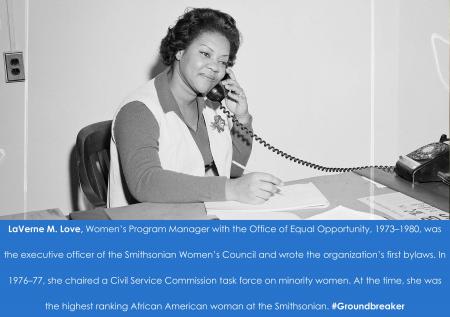 Black and white photograph of a woman holding a phone to her ear at a desk.