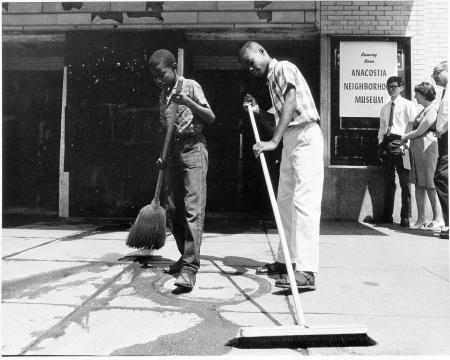 Black and white photograph of two young boys sweeping sidewalk in front of museum entrance.