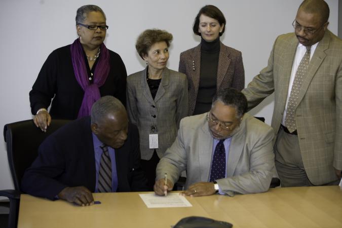 Four people stand and two people sit at a table. One person is signing a document while others look 