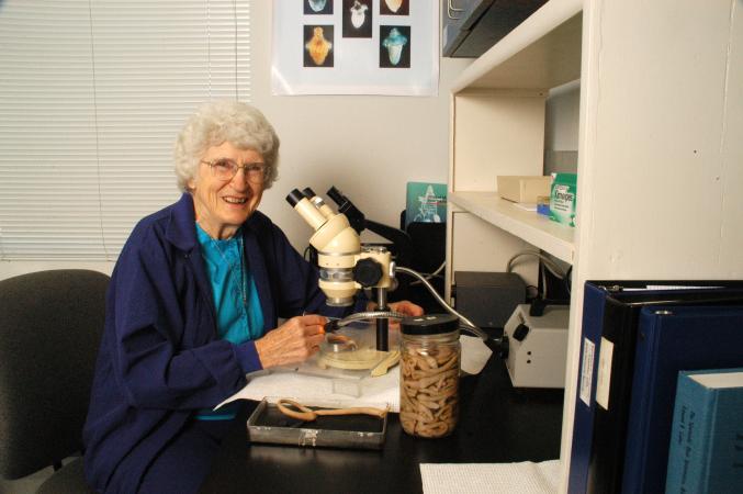 Mary Rice sits at a desk in front of a microscope. A jar of worms is also on the desk.