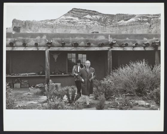 Hirshhorn walks in front of O'Keeffe near a home in the mountains. Hirshhorn is wearing a long coat 