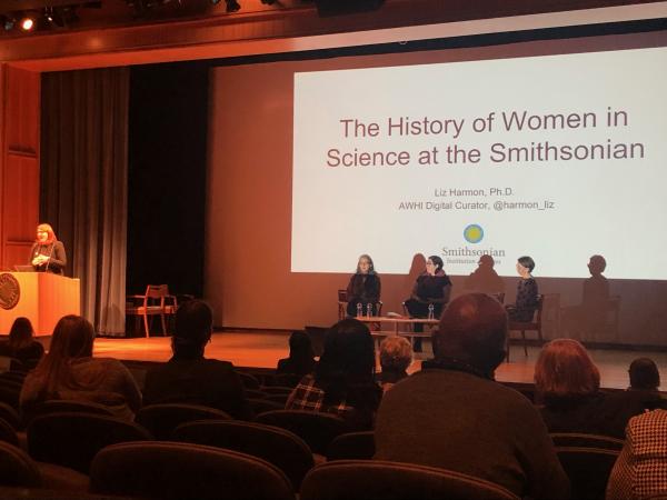 Dr. Liz Harmon stands at a podium, while three women sit in chairs on the stage. A talk, titled 