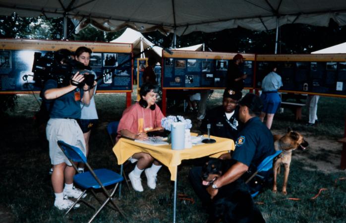 Outside on the National Mall an interviewer sits across a table from two men with two dogs. Recordin