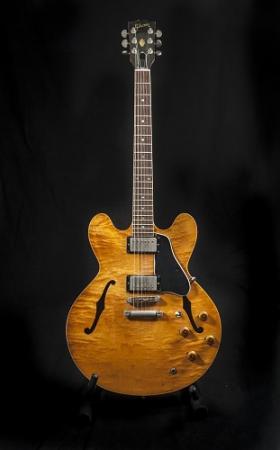 A six-string, semi-acoustic guitar has a light brown maple body photographed on a black background. 