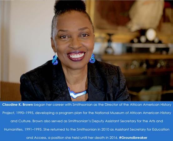 A woman smiles directly at the camera. Her hair is in a bun and she is wearing blue earrings and a n