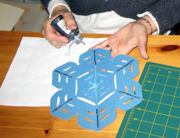 Someone decorates a blue snowflake with glitter glue.