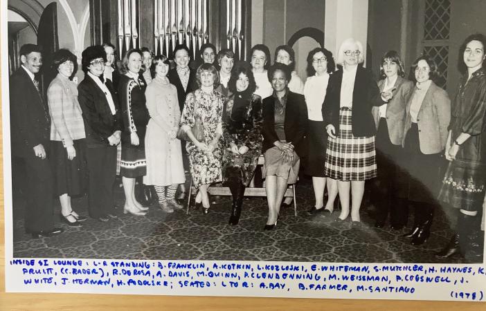 A group of women and one man sit and stand in a room.