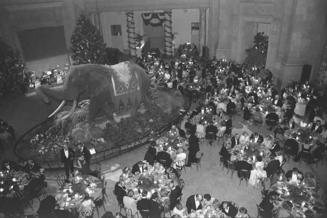 Photograph of men and women eating at round tables, taken from above. A large taxidermied elephant i