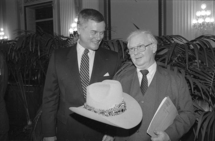 Larry Hagman holds up a "cowboy" hat and stand next to another man. 