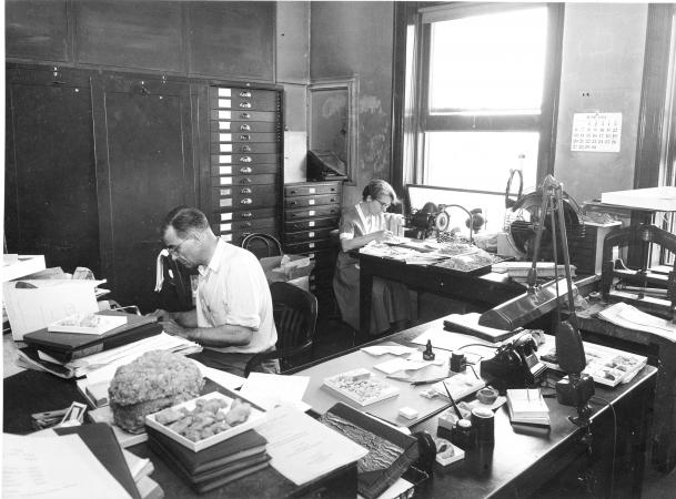 A man and woman sit at work at two long, cluttered desks. Both are wearing glasses. Both seem to be 