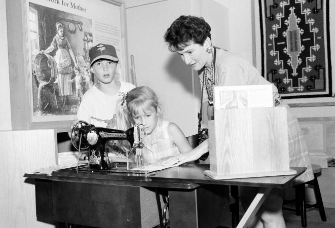 A woman and two children work with a sewing machine on a table.