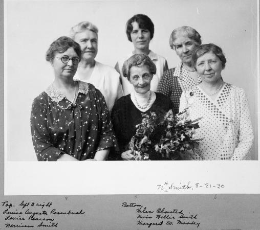 A group of five women stand around an elderly woman holding a bouquet of flowers who is retiring. 