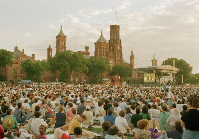 A crowd sits on the Mall facing a stage in front of the Smithsonian Institution Building. 