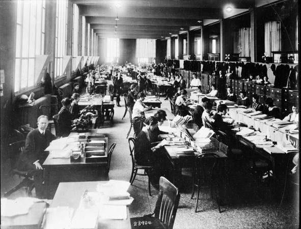 Men and women sit closely at desks that hold piles of paper. 