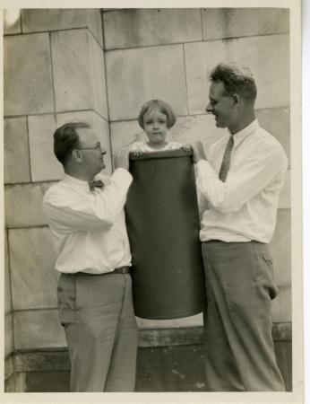 Two men hold a young girl up in a trash can.The girl is looking directly at the camera. 
