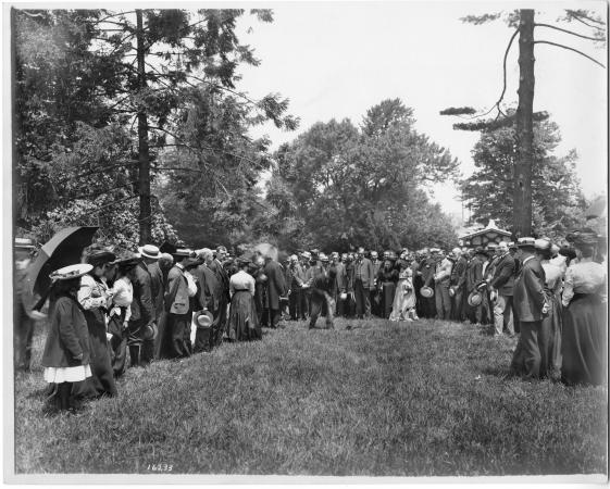 Men and women in nice clothes stand around a field. A man is standing at the center and digging into