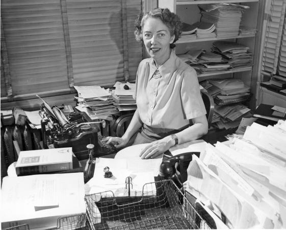 A woman sits at a desk near a typewriter and many stacks of papers. 