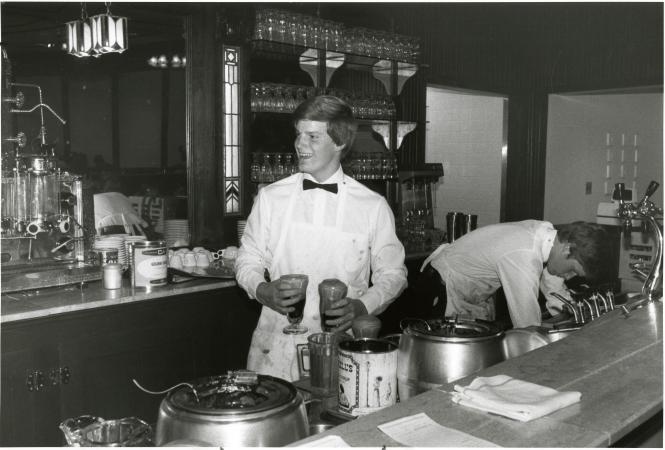 Two men stand behind a counter. One man is holding an ice cream float in each hand.