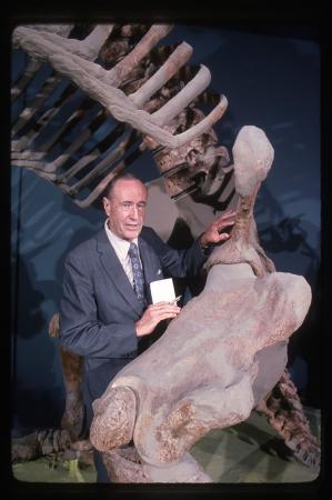 A man in a suit stands next to skeletal bones that are much larger than him. 