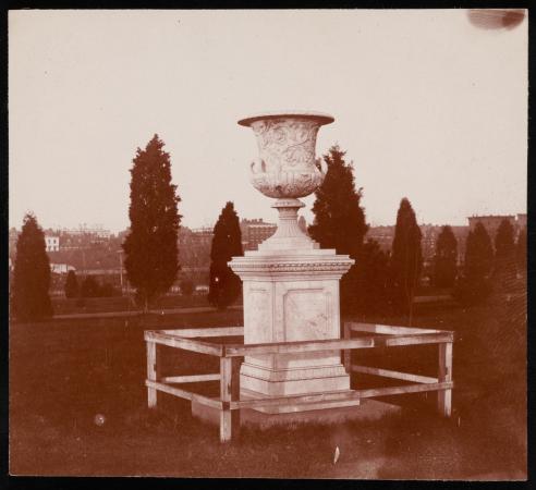 An urn sits in a grassy area. It sits on a pedestal. The base is surrounded by a wood fence. 