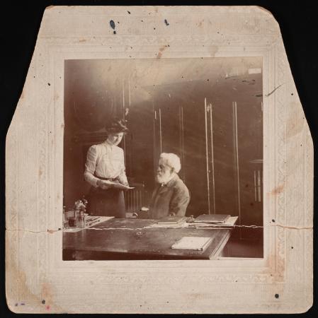Munroe stands over Rhees, who is seated at a desk. She is holding a paper. 