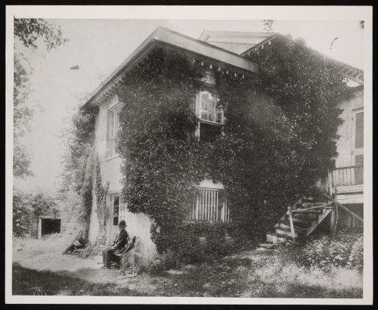 A man sits outside on a bench in front of Holt House. Overgrown greens cover the building. 