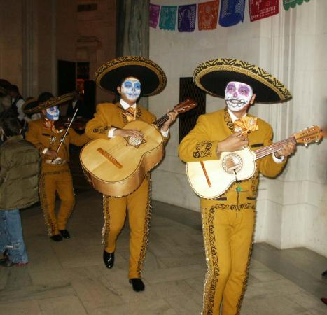 Three men in mariachi outfits hold instruments and walk. They are wearing hats and their faces are p