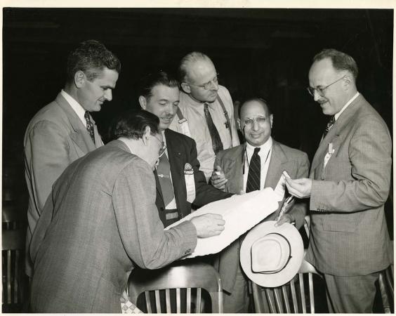 Six men in suits huddle around a square, flat object. One man looks directly at the camera. 