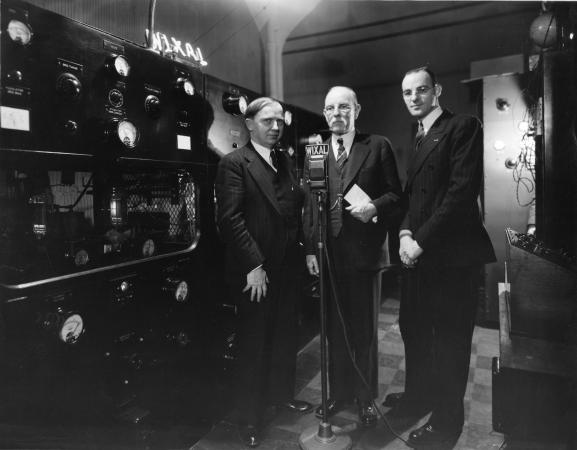 Three men in suits standing behind a microphone and in front of a wall of speakers and meters.