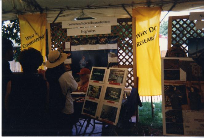 An older man sits at a booth as people wait in line. Banners are visible. They read: Why do research