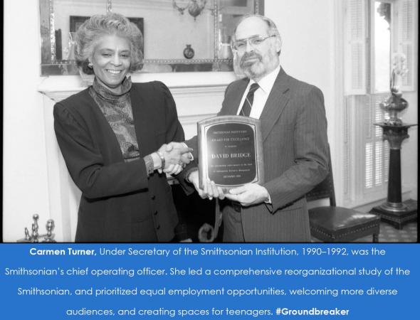 A woman shakes hands with a man who is holding up a plaque. Both are smiling at the camera. 