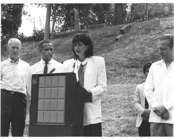 Elena Lombardo stand at a podium outdoors giving a presentation. She surrounded by three men also on